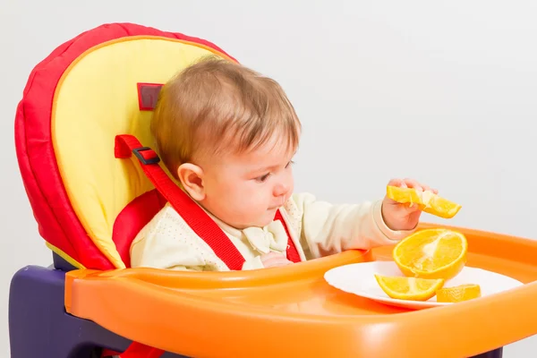 Baby zit in kinderstoel met sinaasappel. — Stockfoto