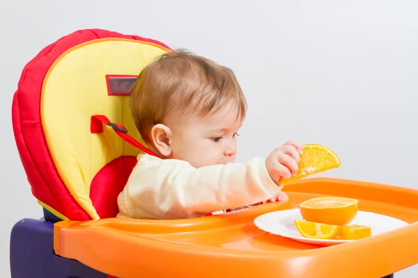 Baby zit in kinderstoel met sinaasappel. — Stockfoto