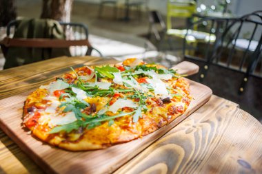 Sebze ve peynirle kaplı yeni pişmiş bir pizza güneşli bir günde, sıcacık bir kafede, ahşap bir tahtanın üzerinde oturur..
