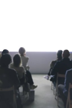 Seyirciler, modern bir mekandaki konferans sırasında parlak bir odada sunum izliyorlar.