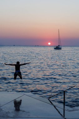 Bir insan gün batımında okyanusa atlarken yakınlarda bir yelkenli yüzer ve yaz neşesi ve özgürlüğünü yakalar.