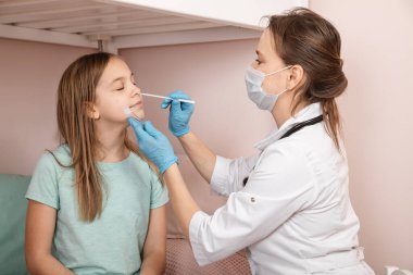 Pediatrician taking nasal mucus test sample from elementary age girl's nose performing respiratory virus testing procedure at home during coronavirus COVID-19 pandemic quarantine