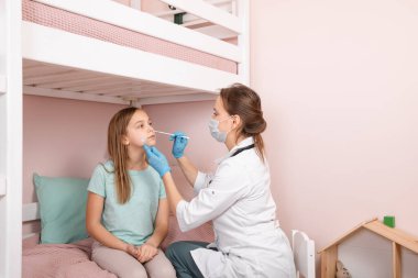 Pediatrician taking nasal mucus test sample from elementary age girl's nose performing respiratory virus testing procedure at home during coronavirus COVID-19 pandemic quarantine