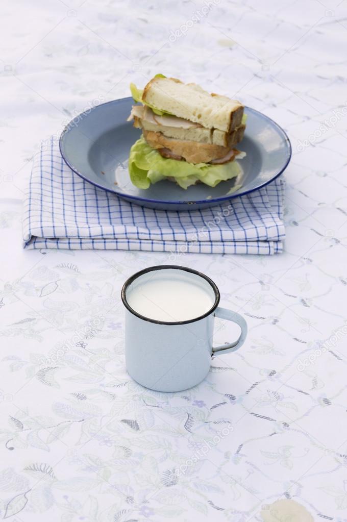 Tin cup of milk with sandwich on plate in background