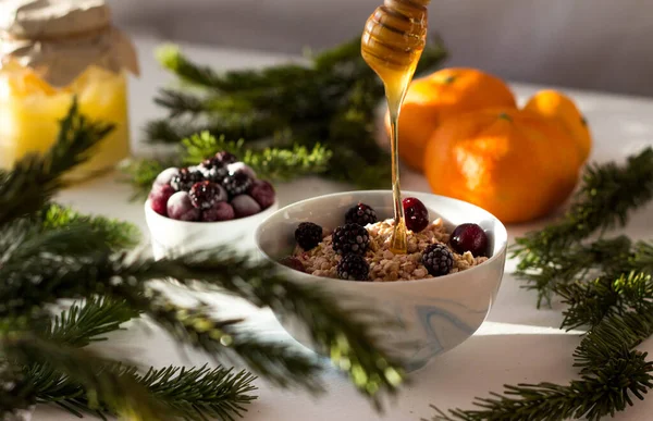 muesli in a white plate on the table, in which honey flows from a spoon on top. Frozen berries lie on top of the muesli, around the fir branches