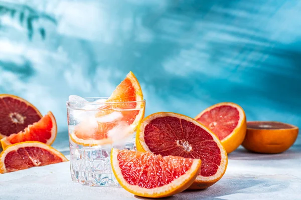 refreshing cocktail with grapefruit on a blue background. a glass glass with ice and grapefruit slices sits on the table among the sliced citrus fruits. background of the shadow of palm trees