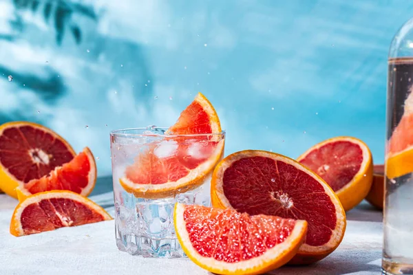 refreshing cocktail with grapefruit on a blue background. a glass glass with ice and grapefruit slices sits on the table among the sliced citrus fruits. background of the shadow of palm trees