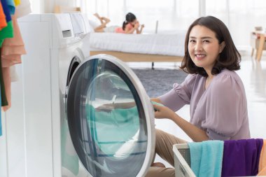 Gülümseyen Asyalı kadın, rutin ev işleri için çamaşırlarını çamaşır makinesine yüklerken yatakta uzanmış, yumuşak bir arka planı olan genç bir kız..