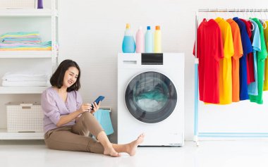 Gülümseyen Asyalı kadın çamaşır odasında çamaşır makinesinin çamaşır yıkamasını beklerken kendini rahatlamış hissederek yerde oturmuş akıllı telefonu izliyor.