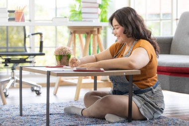 Genç, gülümseyen Asyalı şişman bir kadının portresi. Bir evde oturmuş, bir masada ders çalışıyor ve güzel şeyler yazıyor. Ev konseptinden çalış. Tombul bir kadın yüzüne odaklan..