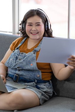 Şişman Asyalı iş kadını kulaklık takıyor, kulaklık takıyor, gülümsüyor ve kucağında bir dizüstü bilgisayarla elinde bir fotoğraf makinesiyle evde gündüzleri çalışan bir kanepede oturuyor..