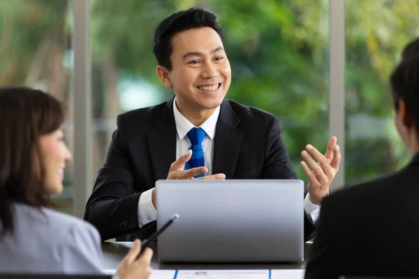 Genç yakışıklı Asyalı işadamının portresi ofisteki şirket işleriyle ilgili bir tartışma sırasında gülümseyip ekibiyle konuşuyor. Ortada siyah takım giyen bir adama odaklan..