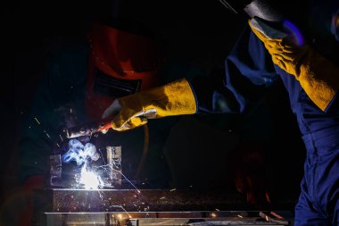İş arkadaşı kaynak yaparken gece vakti fabrikada yeşil mekanik tulum ve güvenlik kaskı takan bir fabrika işçisi....