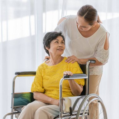 Asyalı orta yaşlı kadın Carre 'ı alıp tekerlekli sandalyedeki yaşlı ve mutlu yüzlü hastaya yardım ediyor..