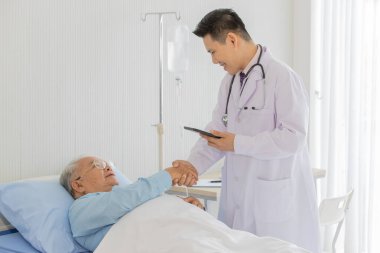 Siyah saçlı, beyaz önlüklü ve steteskoplu genç bir Asyalı doktor sol elinde siyah tablet tutuyordu ve yaşlı şişman Asyalı gri saçlı hasta elini hastane yatağında tutuyordu..