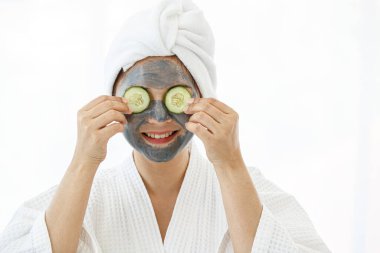 Güzel bir Asyalı kadın spa elbisesi giyip başını beyaz bir havluyla örtmüş ve yüzüne krem sürmüş. Mutluluk dolu salatalıklarla oynuyormuş..