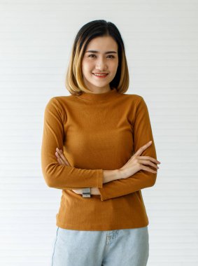 Uzun kollu, kahverengi elbiseli ve kot pantolonlu, beyaz arkaplan ile izole edilmiş şirin gülümseyen Asyalı kadının orta boy portresi.