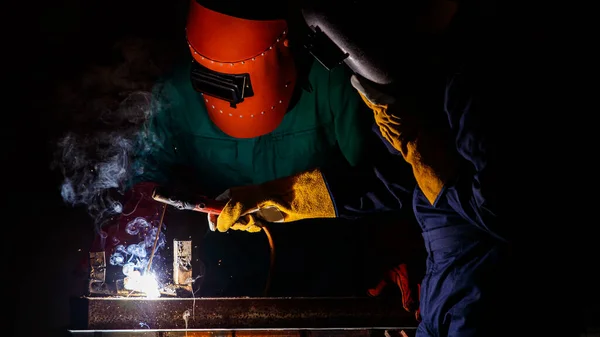 A factory worker wearing a green mechanic coveralls and safety helmet welding metalwork at night time in a factory while his colleague looking at a spark from welding.