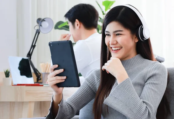 Young Asian black long hair woman staying in living room wearing headphone and holding tablet computer and widely smile to gadget screen while man working blur in blackground.