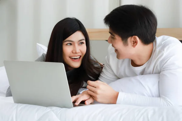 若いアジアの黒い長い髪の女性と明確な顔の男の恋人がベッドの上に横たわり リラックスして幸せと一緒にインターネットを提供するためにノートパソコンを使用して — ストック写真