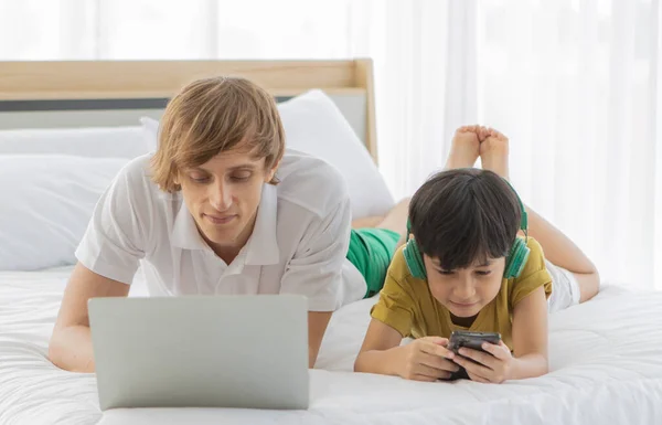 白人の父と混合レースの息子はベッドの上に横たわって デバイスとインターネットに接続します 父はノートパソコンを使い子供はスマートフォンを使います — ストック写真