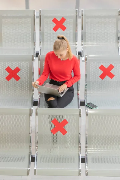 Female tourist sitting on row seats in airport lobby chair with social distancing sign and using note computer while waiting for flight chaning. New normal journey concept.