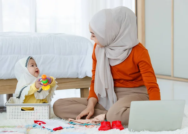 一位可爱而美丽的亚洲穆斯林母亲在笔记本电脑上做着在线推销员的工作 同时照顾着她的小女儿 — 图库照片