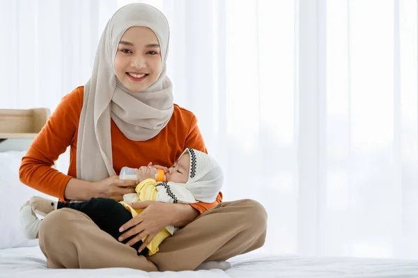 一位可爱美丽的亚洲穆斯林母亲坐在床上 怀里抱着熟睡的女婴 用奶瓶喂她 — 图库照片