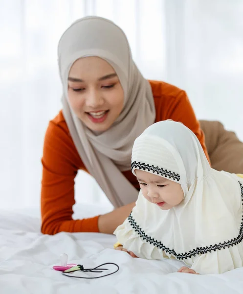 一位漂亮可爱的亚洲穆斯林 身穿头巾裙 与女儿玩耍 与床上的奶嘴玩耍 — 图库照片