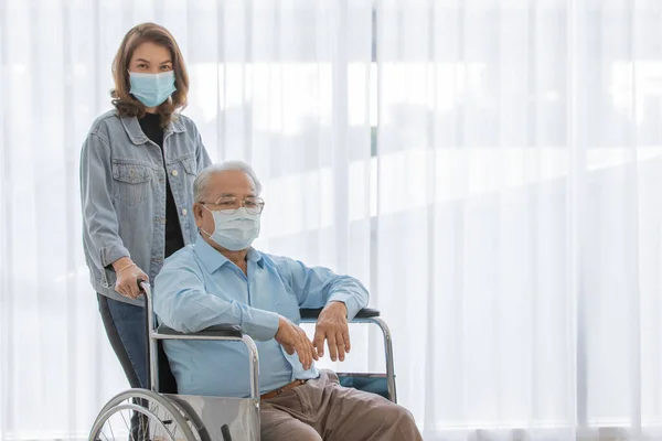 An old fat asian male patient with gray hair wearing light blue shirt and brown pants sitting on wheelchair when his daughter wearing jeans shirt stand holding the handles look at camera together.