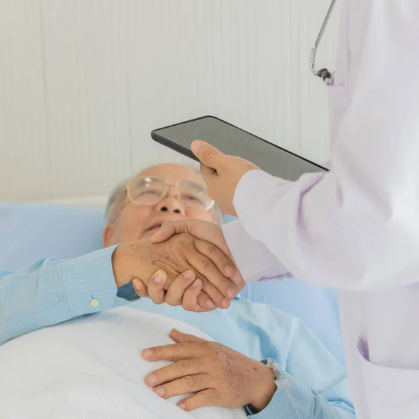 Close up shot of holding hands of old asian fat patient lay down in hospital bed and doctor wearing white lab coat with stethoscope and black tablet.