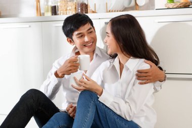 Beyaz gömlek ve kot pantolon giyen genç ve çekici Asyalı çift ellerinde beyaz kahve kupalarıyla beyaz mutfakta oturuyor ve birbirlerine gülümsüyorlar. Sağlıklı ve mutlu aşk ve yemek yapma kavramı.