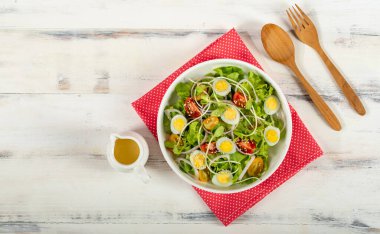 Büyük beyaz bir kase marul, haşlanmış yumurta, soğan ve domatesli filiz salatası kırmızı puantiyeli kumaş üzerine susam sosu beyaz ahşap masa. Yanında küçük zeytinyağı kavanozu..