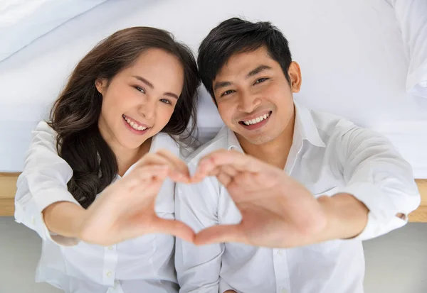 若い幸せな魅力的なアジアのカップルのきれいな白いシャツを一緒にベッドルームの床の上にカメラを手の心のサインを行う上で座って着ている 愛と幸せな関係の概念 — ストック写真