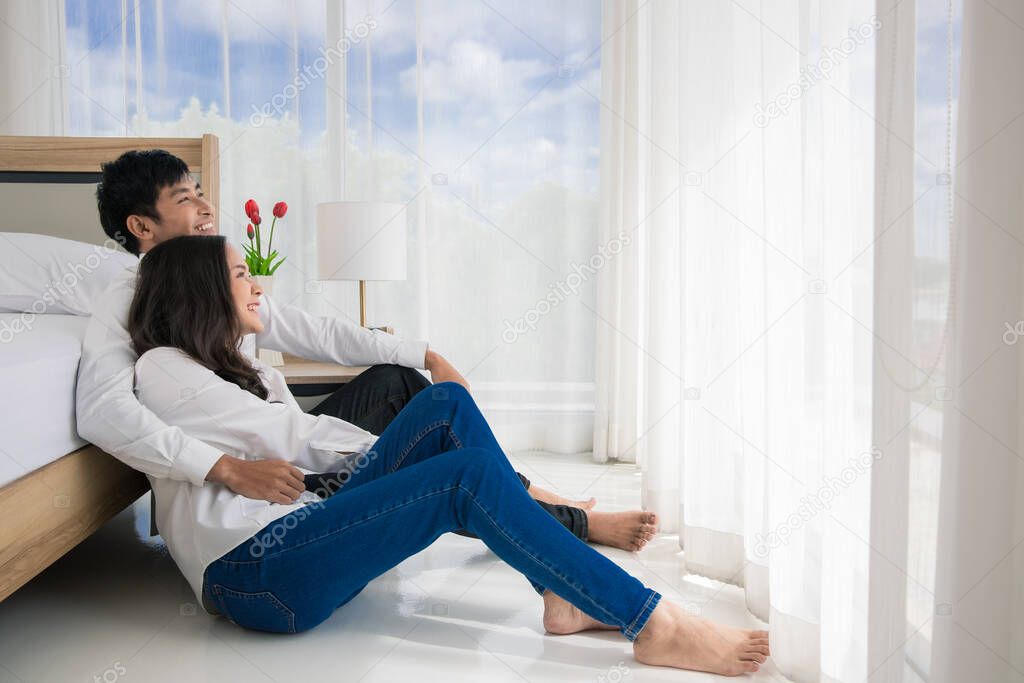 Young happy attractive Asian couple hugging sitting together on the floor in bedroom looking at blue sky view outside window through white curtain. Concept for love and happy relationship.