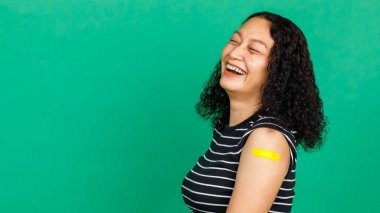 Orta yaşlı Asyalı kadın kolunu bandajlı göstererek gülüyor. Yeşil arka planda Covid 19 virüsü aşısı olduğunu gösteriyor. Covid 19 aşısı için kavram.