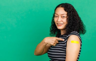 Asyalı orta yaşlı, gülümseyen ve koluna bandaj bandıyla bakan bir kadın yeşil arka planda Covid 19 virüsü aşısı olduğunu gösteriyor. Covid 19 aşısı için kavram.