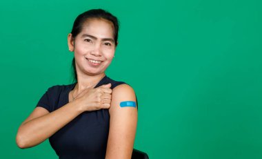 Orta yaşlı Asyalı kadın kolunu bandaj bandıyla göstererek yeşil arka planda Covid 19 virüsü aşısı olduğunu gösteriyor. Covid 19 aşısı için kavram.