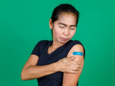 Orta yaşlı Asyalı kadın kolunu bandaj bandıyla göstererek yeşil arka planda Covid 19 virüsü aşısı olduğunu gösteriyor. Covid 19 aşısı için kavram.