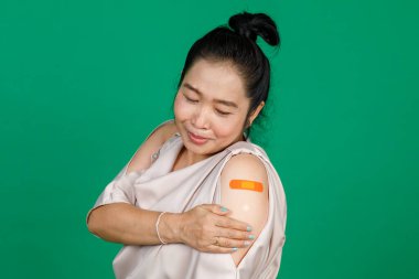 Orta yaşlı Asyalı kadın kolunu bandaj bandıyla göstererek yeşil arka planda Covid 19 virüsü aşısı olduğunu gösteriyor. Covid 19 aşısı için kavram.