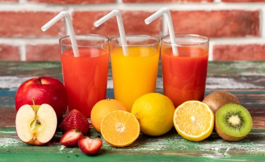 Bardakta, elmada, portakalda ve çilek suyunda samanla farklı meyveler, tuğla duvarın önündeki renkli tahta üzerinde canlandırıcı görünüyor. Taze meyvelerle süsleme.