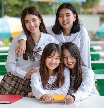 Okulun kampüsünde oturan beyaz tişörtlü, kahverengi kareli okul üniformalı iki çekici Asyalı kız öğrenci. Eğitim kavramı.