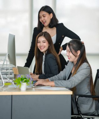 Resmi takım elbiseli üç çekici Asyalı bayan ofis arkadaşı masadaki bilgisayar ve dizüstü bilgisayarla gülüyor ve birlikte çalışıyorlar..