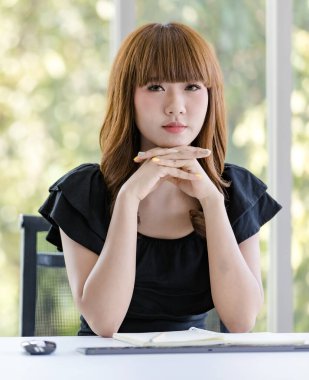 Asyalı iş kadını çenesinin altında el ele tutuşup bilgisayar ekranına ciddi ruh hali, bulanık geçmiş ve ofiste çalışma kavramı ile bakıyor..