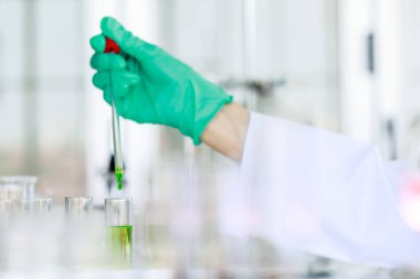 Laboratuvar önlüklü tanınmayan bilim adamı ve pipetli eldiven laboratuarda kimyasal deney yaparken yeşil sıvıyı şişeye döküyor.