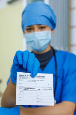 Hastaya doktor tarafından gönderilen Covid-19 aşı kayıt kartı pasaport kağıdının yakın plan fotoğrafı. Mavi hastane üniforması, plastik eldiven ve bulanık arka plan steteskobu takıyor..