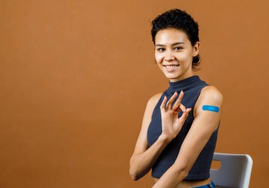 Coronavirus Covid-19 aşısı vurulduktan sonra omuzundaki mavi alçıda el işaretini gösteren gülümseyen Asyalı, neşeli ve seksi kadın hastanın yakın çekim portresi..