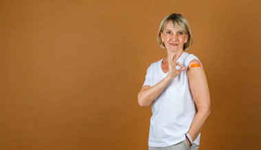 Kafkas kıdemli sarışın kadının portresi. Kamera önünde el işareti ve Coronavirus aşısı olduktan sonra omzunda turuncu bandaj var..
