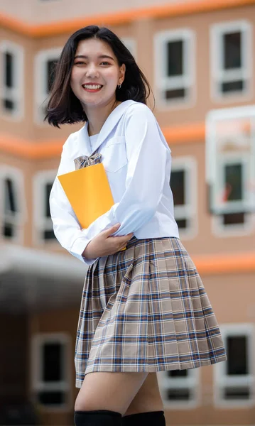 Portrait of young attractive female asian high school students in white shirt brown checkered school uniform holding yellow document files in school campus. Concept for education.