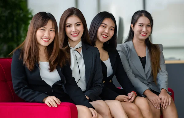 4つの魅力的なアジアの女性のオフィスの同僚のグループは 正式なビジネススーツの赤いソファの上にオフィスのカメラで笑顔に座っている 近代的な事務所業務の概念 — ストック写真